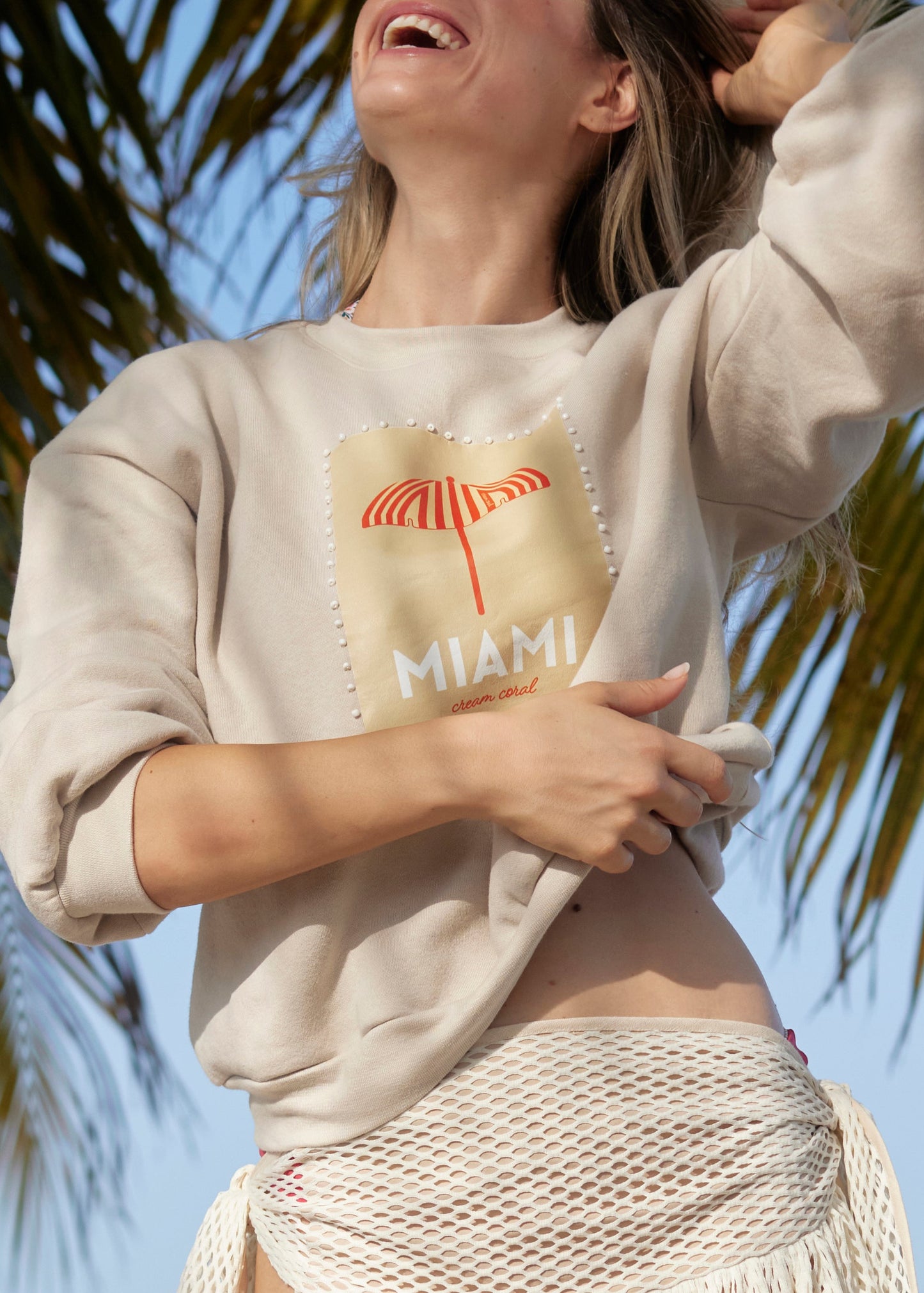 Woman wearing a beige sweatshirt with a 'Miami' design, standing against a palm tree background.