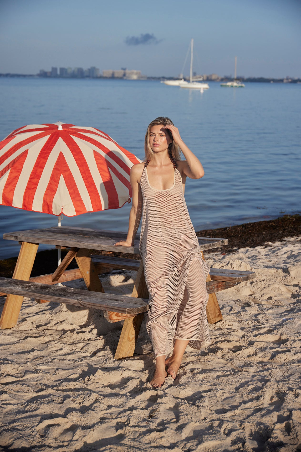 Woman in a beige dress standing on a sandy beach with a red and white striped umbrella and picnic table.