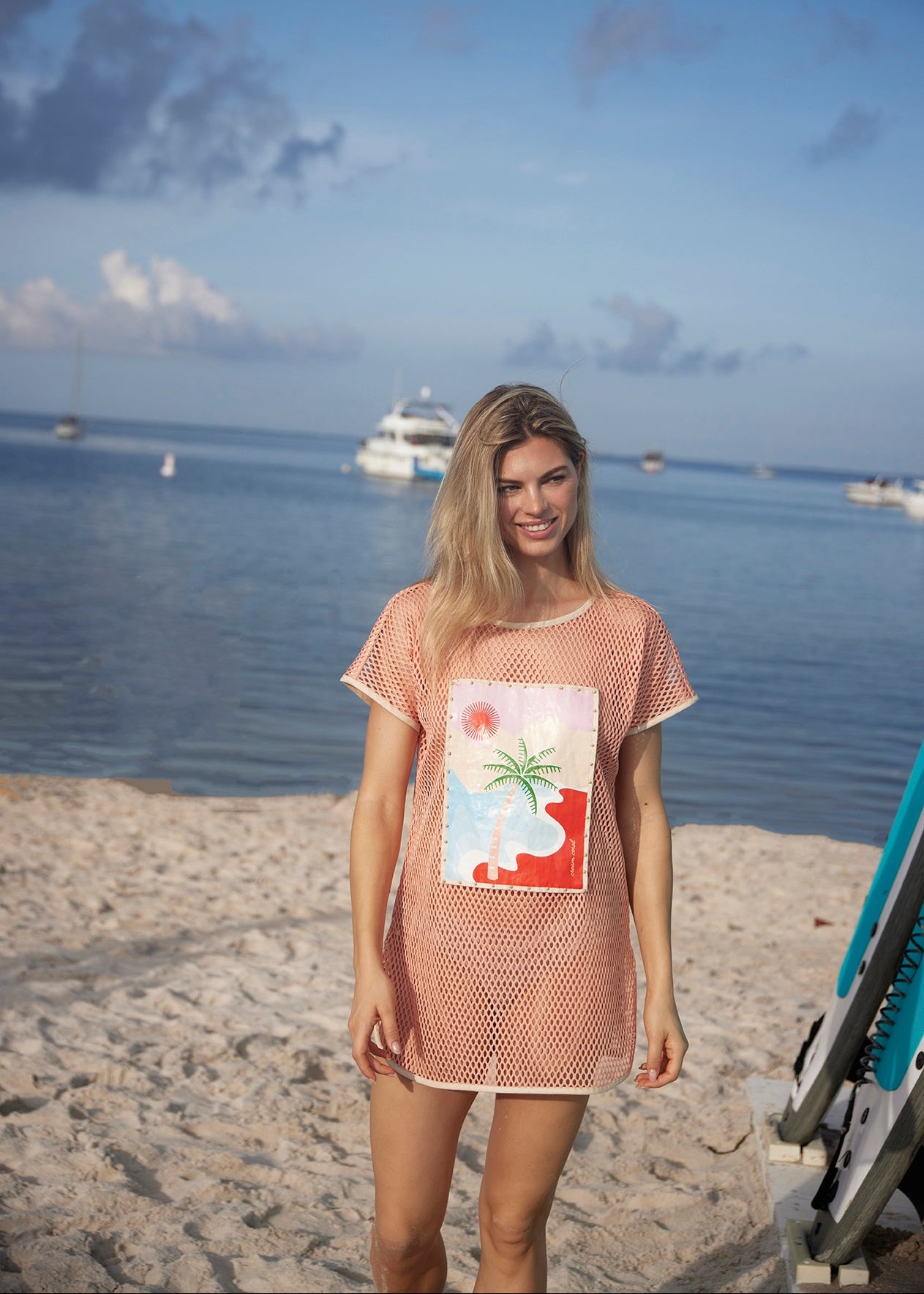 Woman in a beach dress standing on a sandy beach with water and boats in the background